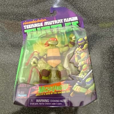 Nickelodeon Teenage Mutant Ninja Turtles Michelangelo action figure. The packaging describes Michelangelo as the "JOKESTER & HARD-HITTING NUNCHUCK HERO".  It includes weapon accessories, such as his iconic nunchaku(B8)
