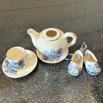 Miniature porcelain dollhouse tea set with a blue floral pattern, including a teapot and a teacup with saucers, and a pair of decorative baby shoes,the items appear to be part of the Royale Stratford Country Cottage (B12)