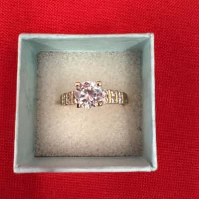  Women's Gold Tone Ring featuring a Prominent Round Center Stone & Channel Set Accents Along the Band
